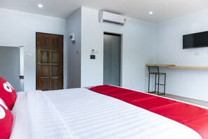 Habitación de hotel con una cama, puerta de madera, unidad de aire acondicionado, televisor montado en la pared y una pequeña mesa con una silla.