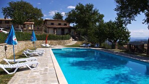 A swimming pool with lounge chairs and umbrellas, surrounded by stone walls and trees, with buildings in the background. - Agriturismo Casanova (Gubbio)