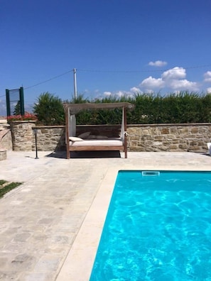 A pool area with a stone wall, a wooden swing chair, and a clear blue pool.