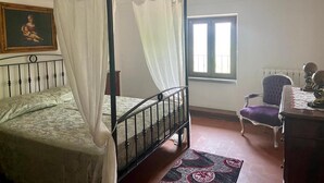A four-poster bed with white drapes, a painting above the bed, a window with blinds, a chair, and a table with a decorative item. - Agriturismo Casanova (Gubbio)