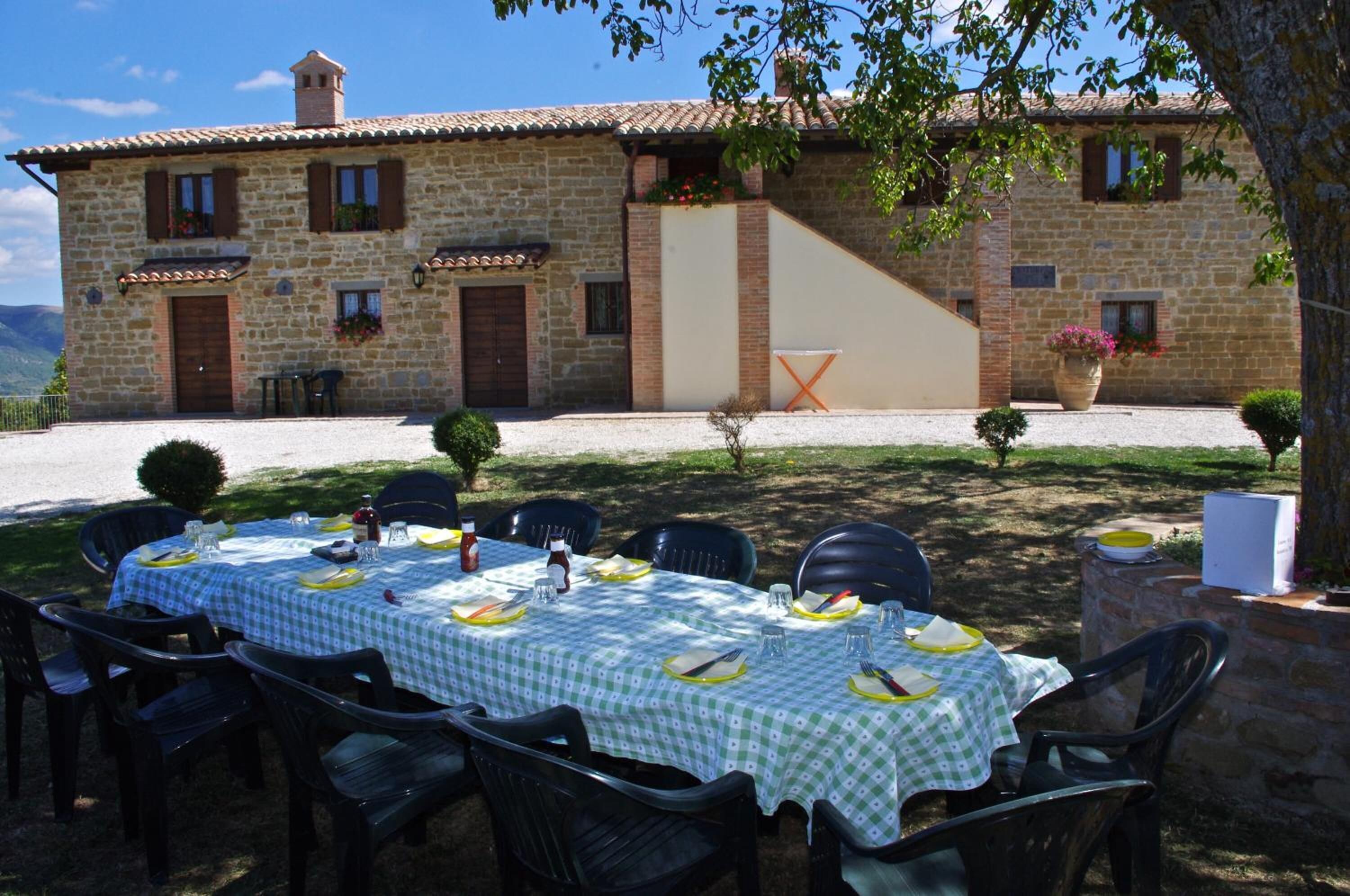 An outdoor dining area with a stone building in the background, a table set for four, and a tree providing shade.
