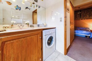 A bathroom with a washing machine, wooden cabinets, a mirror, and a bed in the adjacent room. - Homerez - Apartment in Bolquère, mountain view (Bolquère)
