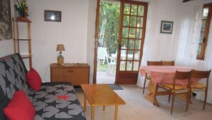 Een woonkamer met een bank, een houten tafel en een eettafel met stoelen. Er is een deur die leidt naar een buitenruimte met stoelen.