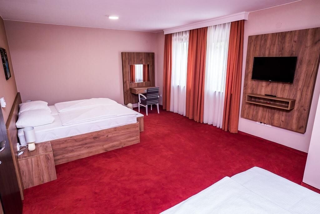 Habitación de hotel con una cama de madera, un escritorio, una silla, un televisor montado en la pared y una alfombra roja.