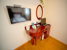 Habitación de hotel con televisor de pantalla plana montado en la pared, un escritorio de madera con una computadora portátil, un hervidor y algunos artículos de aseo.