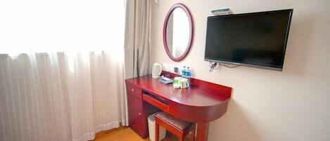 A hotel room with a wooden desk, a mirror, a flat-screen TV, and a window with curtains.