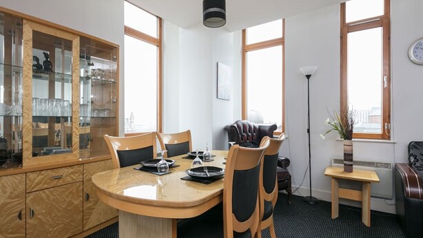 A modern kitchen with a round dining table, wooden chairs, and a glass cabinet with kitchenware. - My-Places City Living Apartment 06 (Manchester)