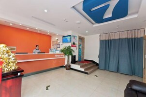 A hotel reception area with a receptionist, a reception desk, a large number '7' sign, a potted plant, and a blue curtain.