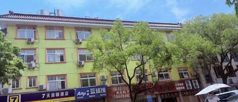 A multi-story building with several ground-floor shops. There are trees in front of the building, and a person is riding a motorcycle with an umbrella.