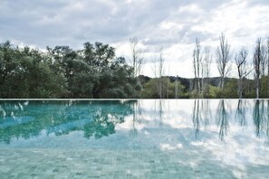 Une piscine aux eaux cristallines, dont le fond est recouvert de carreaux, entourée d’arbres et surplombée d’un ciel nuageux.