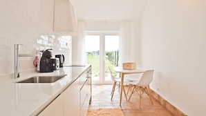A modern kitchen with white cabinetry, a built-in oven, and a coffee machine. There is a dining area with a white table and chairs, and a view of a garden through a glass door. - Herdade do Barrocal de Baixo (Montemor-o-Novo)