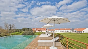A poolside area with lounge chairs and umbrellas, a brick deck, and a view of a building complex in the background.
