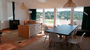 A modern living room with a sofa, dining table, chairs, a fireplace, and large windows offering a view of the outdoors. - Herdade do Barrocal de Baixo (Montemor-o-Novo)