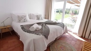 A neatly made bed with a patterned blanket and rolled towels, a bedside lamp, and a view of a garden through a large window. - Herdade do Barrocal de Baixo (Montemor-o-Novo)