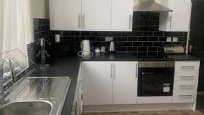 A compact kitchen with white cabinets, a black tiled backsplash, a stainless steel sink, a toaster, and a microwave. - 6 Bedroom Chorlton Townhouse (Manchester)
