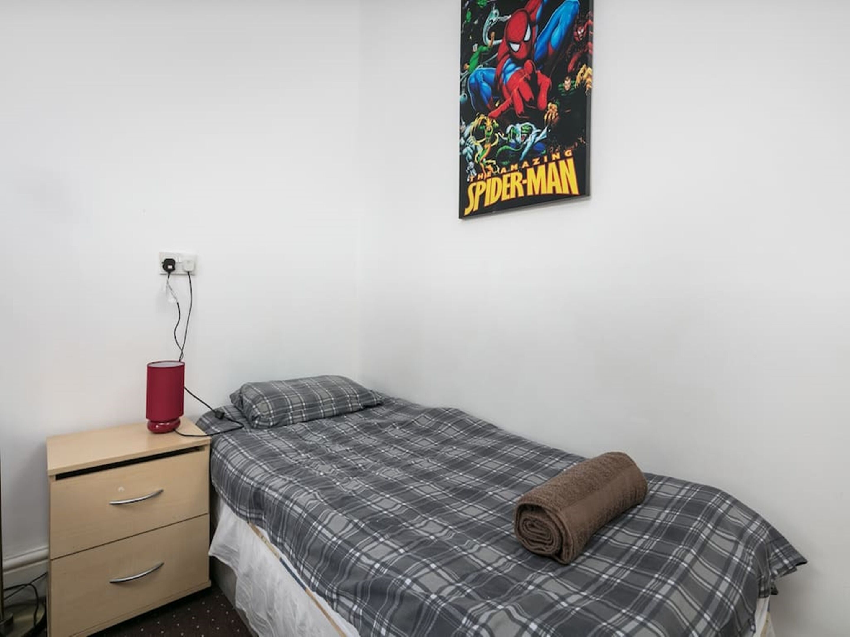 Een slaapkamer met een bed, nachtkastje, lamp en een Spider-Man poster aan de muur.