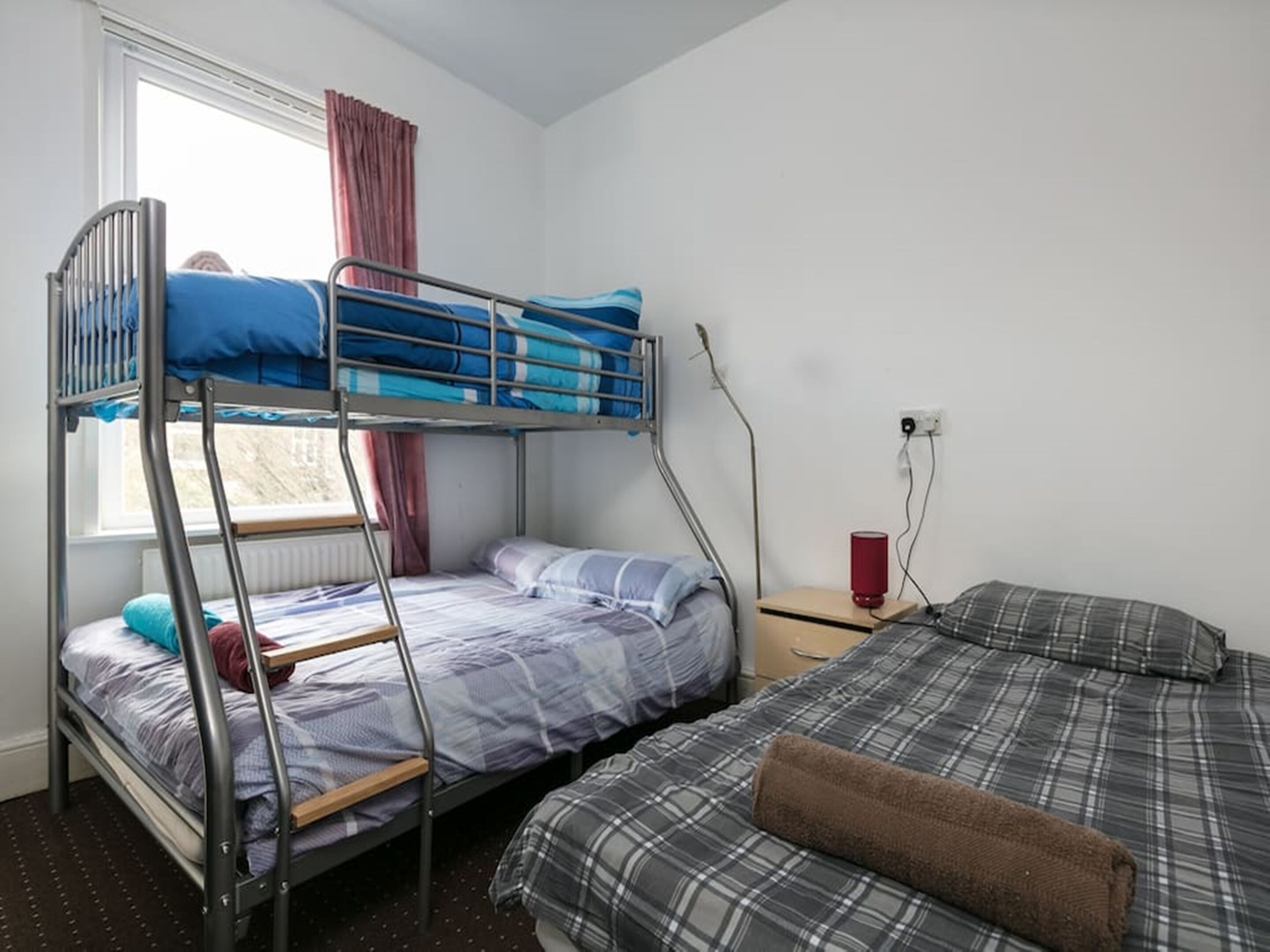 A dormitory room with bunk beds, a window with curtains, a bedside table with a lamp, and a wall socket.