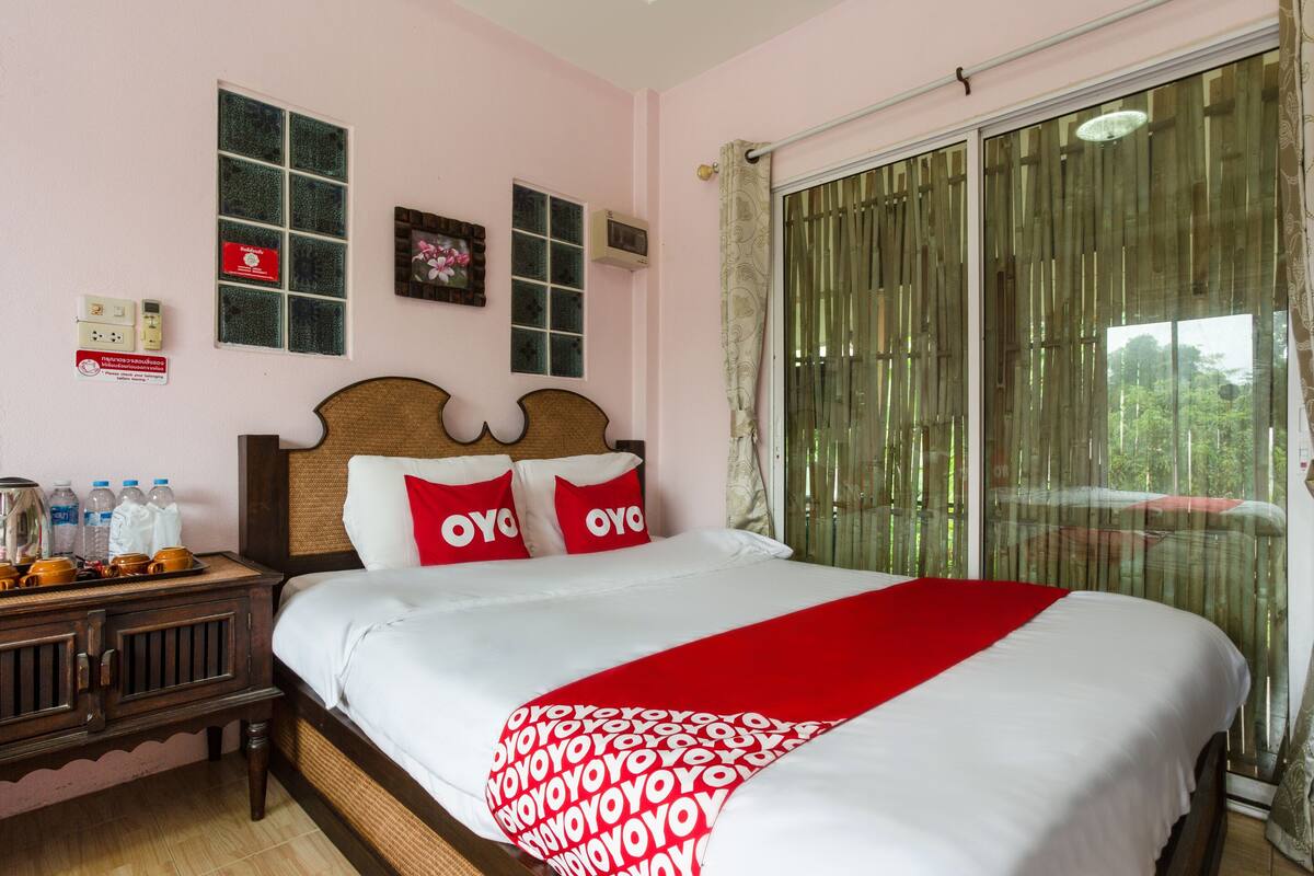 Habitación de hotel con cama, mesita de noche, ventana con cortinas y vistas a áreas verdes.