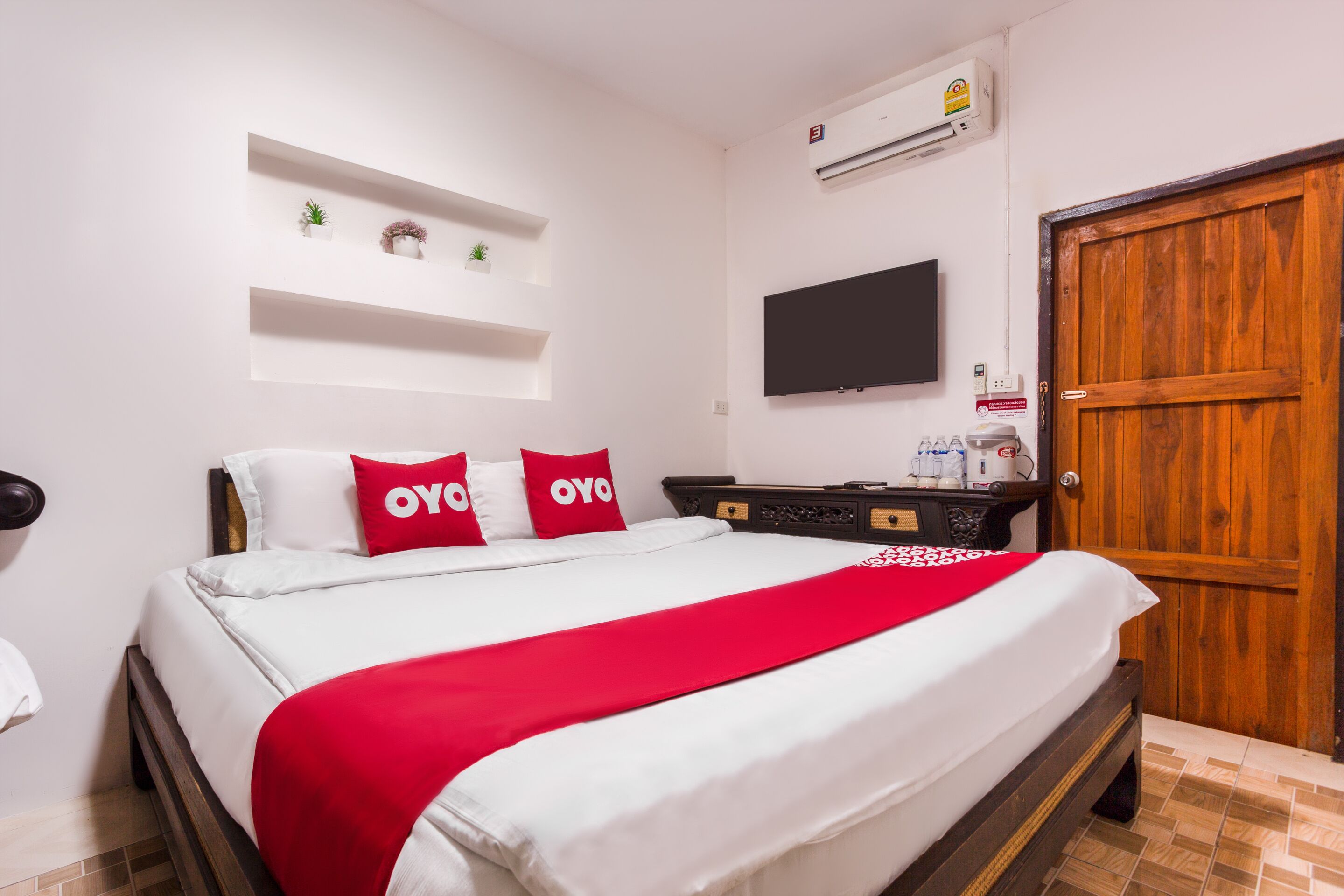 A neatly arranged hotel room with a bed, a wooden wardrobe, a television mounted on the wall, and a small shelf with decorative items.