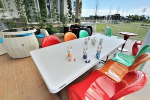 Een terras met een witte tafel, kleurrijke stoelen en uitzicht op een park.