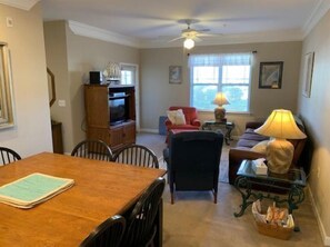 A living room with a dining table, chairs, a sofa, a red armchair, a television on a wooden cabinet, a ceiling fan, and a window with curtains.