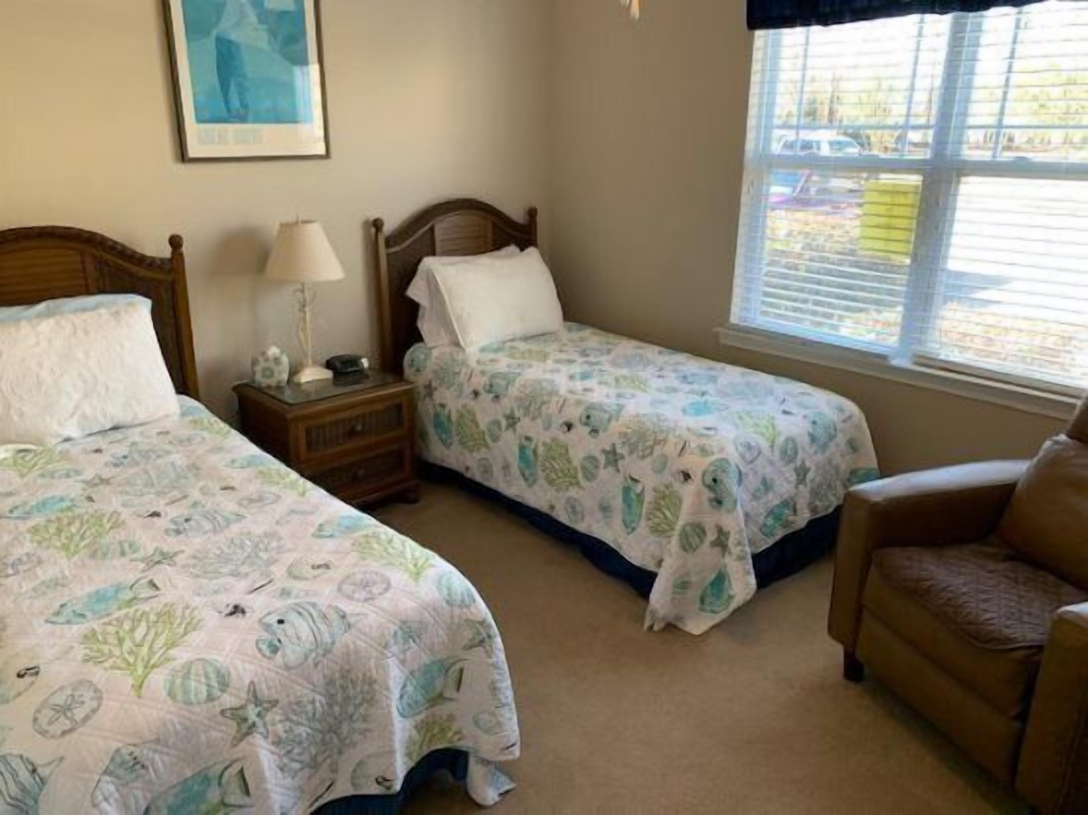 A room with two single beds, a nightstand with a lamp, a chair, and a window with blinds.