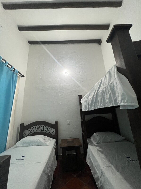 A small, dimly lit room with two single beds, a wooden headboard, a small table, and a blue curtain.