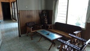 A room with a wooden bench, a small table, and a decorative chair. There is a painting on the wall and a window with curtains. - Duta Karya Hotel (Buleleng)