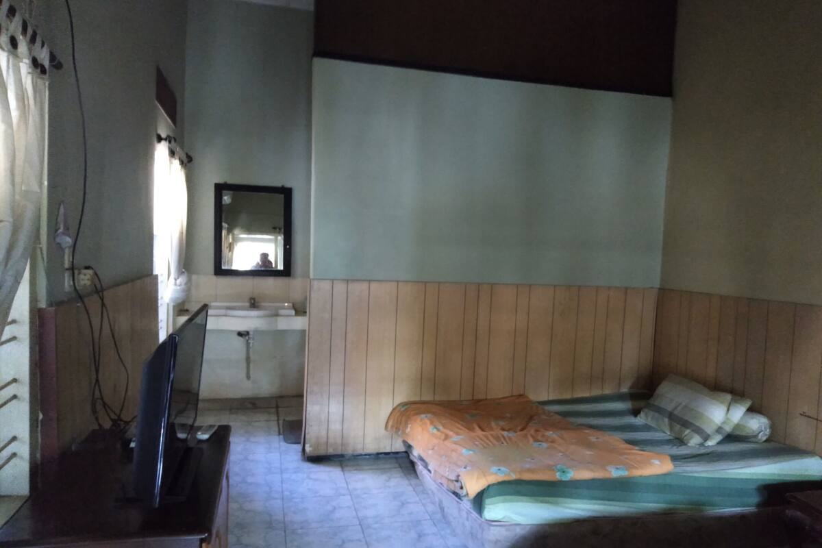 Ein Schlafzimmer mit einem hölzernen Kopfteil, einem Einzelbett mit orange-grüner Decke, einem kleinen Nachttisch und einem Fernseher auf einer Kommode.