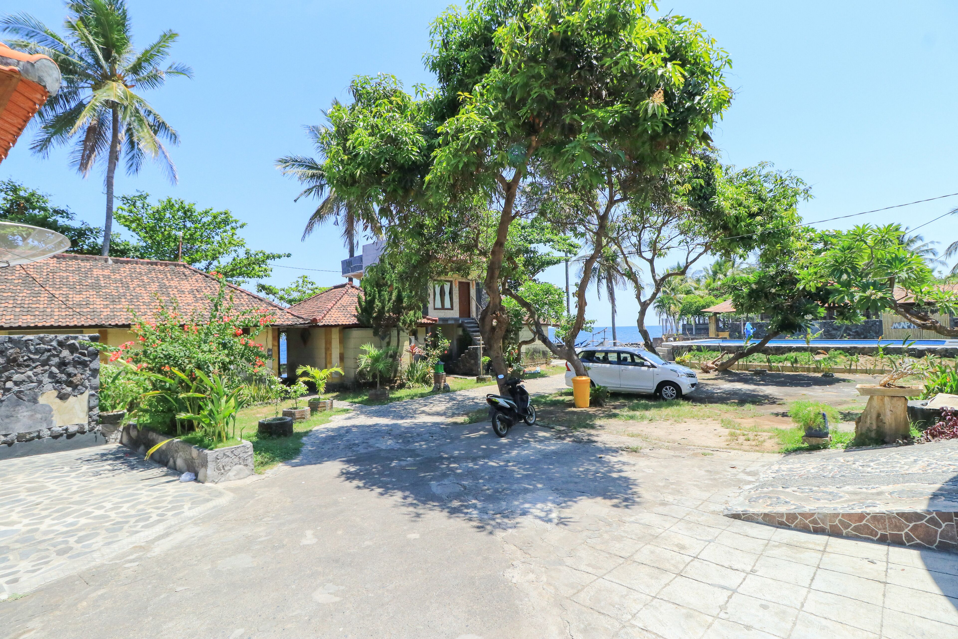 A residential area with a paved driveway, a parked scooter, and a white car. There are trees and a stone wall visible.