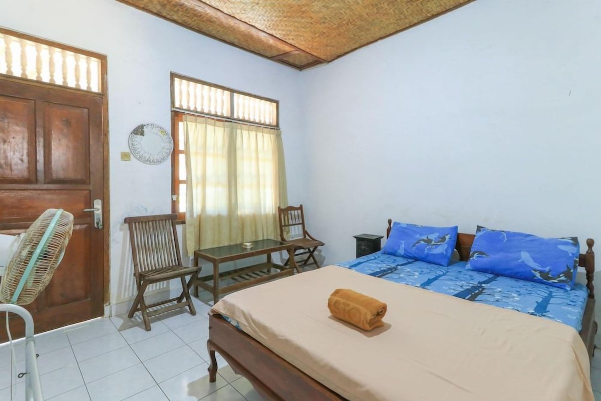A bedroom with a bed, a wooden table, two chairs, a fan, and a window with curtains.