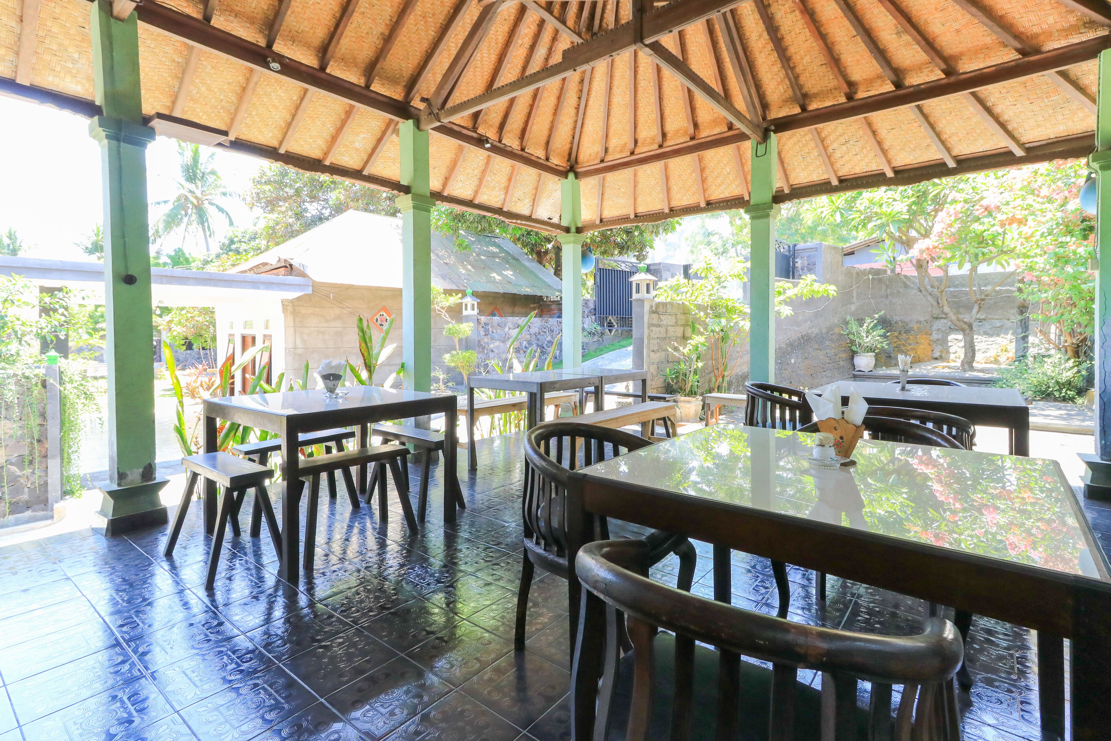 Un área de comedor al aire libre cubierta con mesas y sillas, techo de paja y vistas del jardín circundante y edificios.