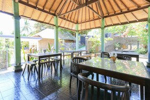 An outdoor covered dining area with tables and chairs, a thatched roof, and a view of the surrounding garden and buildings. - Hotel Tara Bar & Resto Air Sanih (Kubutambahan)