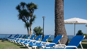 A row of blue and white lounge chairs aligned along a grassy area with a palm tree and a lamp post. - Hotel Restaurante Glasgow (Oia)