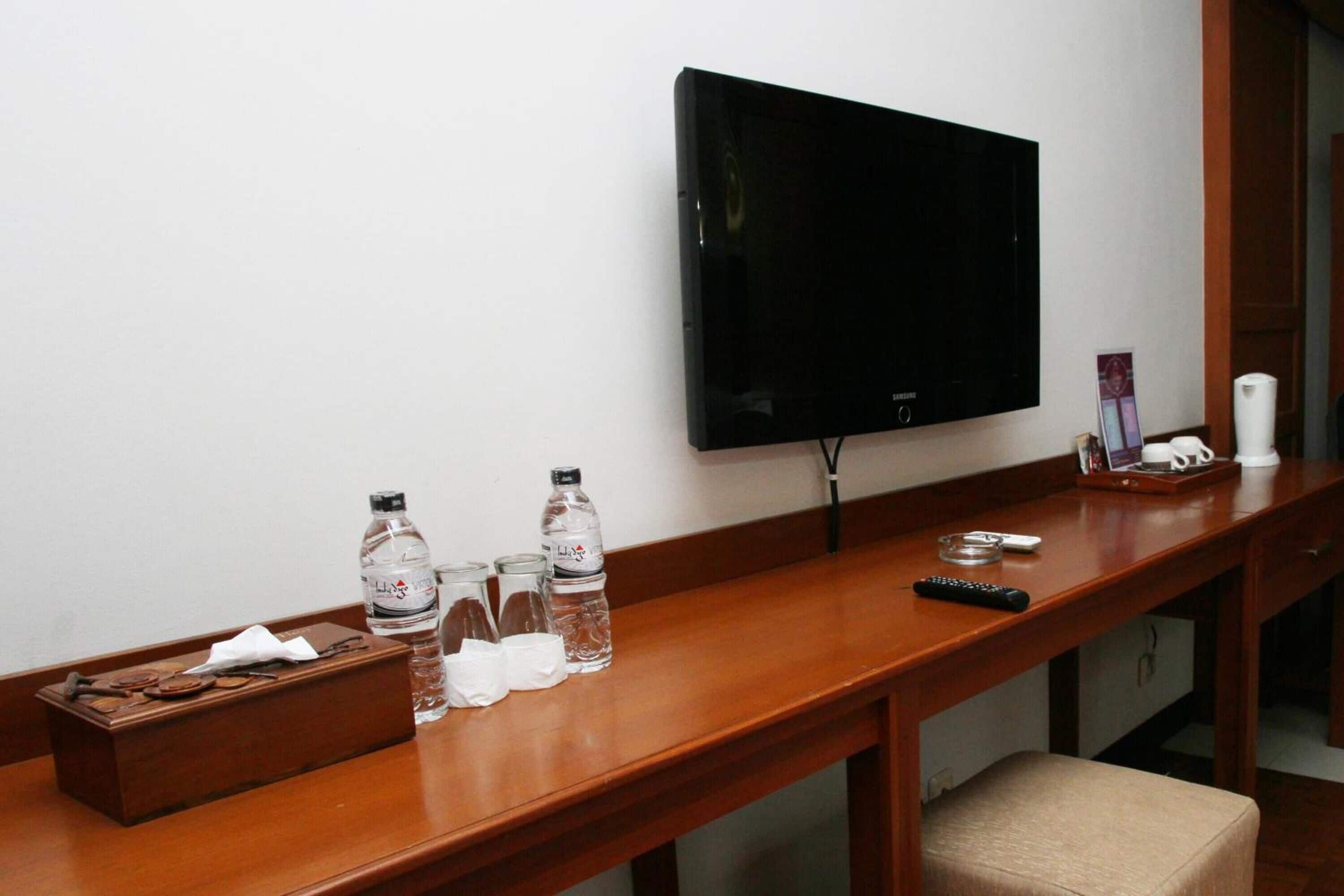 A hotel room with a wooden desk, a flat-screen TV mounted on the wall, a remote control, and a tray with glasses and bottled water.