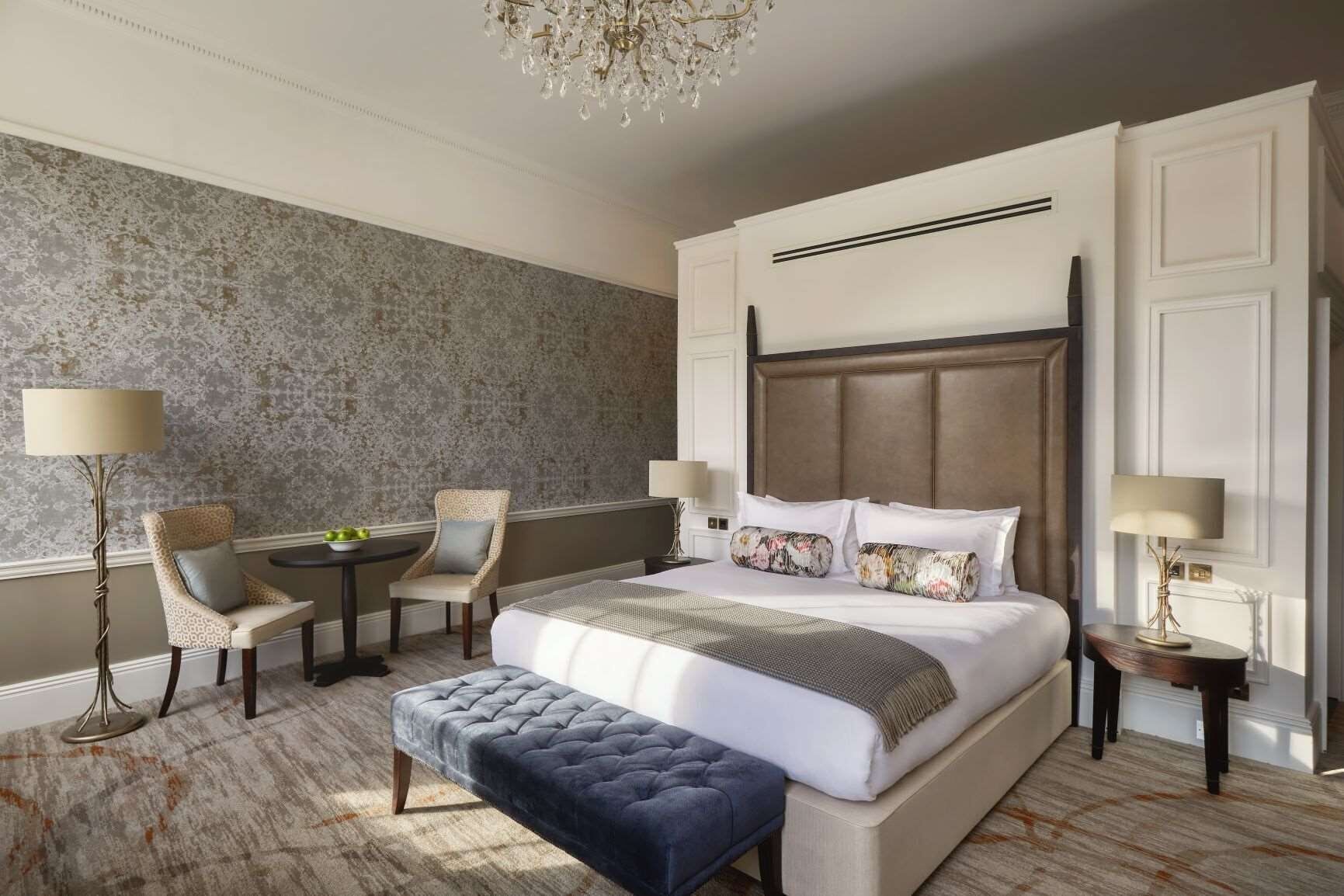 A hotel room with a large bed, two chairs, a small table, a bench, and a chandelier.