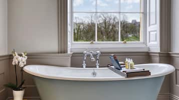 A freestanding bathtub in a bathroom with a window, a potted orchid, and a stack of books.