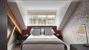 Ein Schlafzimmer mit schrägen Wänden, einem großen Bett mit Kissen, einem Nachttisch, einer Lampe und einem Fenster mit Blick auf Gebäude.