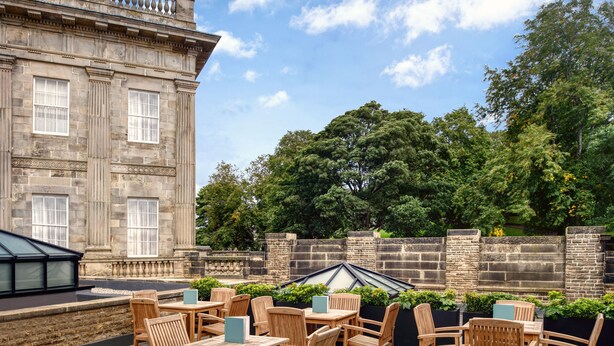 An outdoor patio with wooden furniture and a stone building in the background. - Buxton Crescent (Buxton)