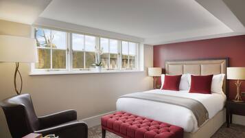 A bedroom with a large bed, a red upholstered bench, a chair, a small table with books and a cup, and a window with a view of a tree and sky.