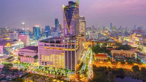 A high-rise hotel with a distinctive triangular top, surrounded by a well-lit urban area with other buildings and a river. - Ratchada 17 Place (Bangkok)