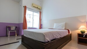 A hotel room with a bed, a chair, an air conditioning unit, a window with curtains, and a nightstand with a lamp.