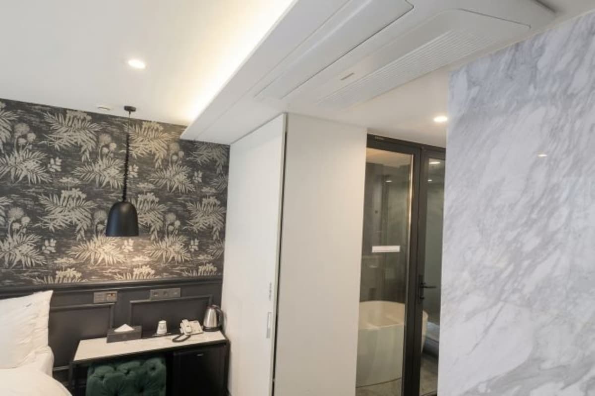 A modern hotel room with a bed, a desk with a telephone, a black pendant light, and a marble wall.