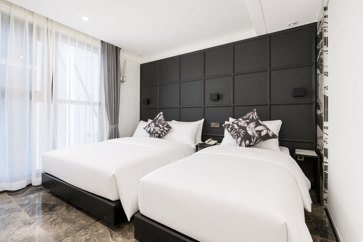 A hotel room with two beds, a dark headboard, a bedside table with a phone, and a large window with sheer curtains.