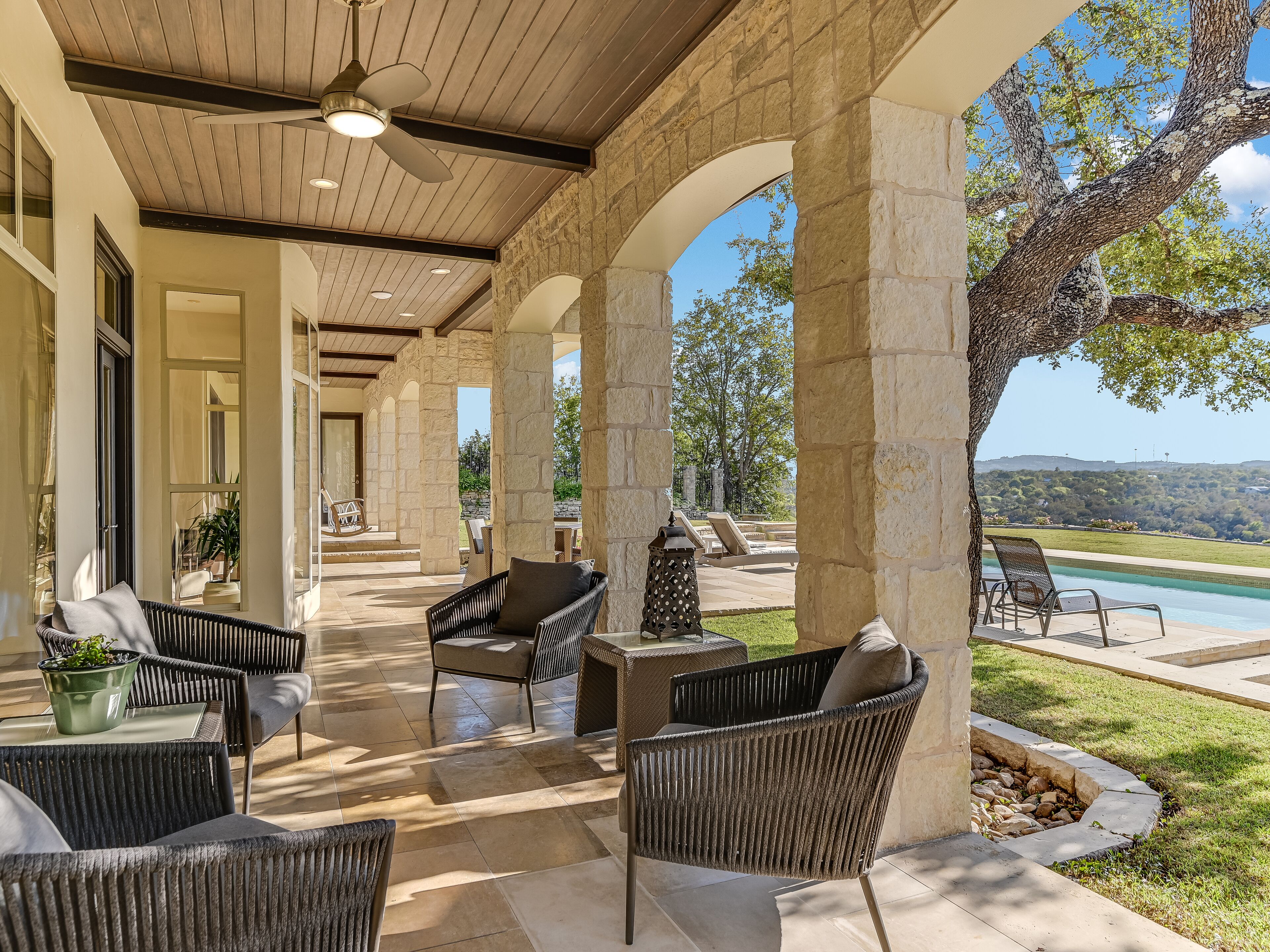 A covered outdoor patio with wicker furniture, stone pillars, and a view of a swimming pool and landscape.