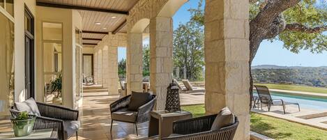 A covered outdoor patio with wicker furniture, stone pillars, and a view of a swimming pool and landscape.