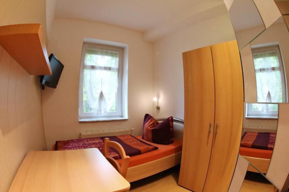 A compact room with a bed, a small table, a wardrobe, and a television mounted on the wall.