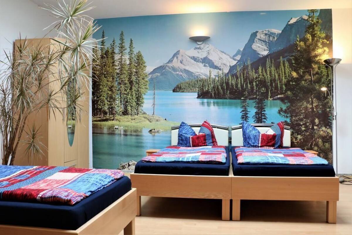 A bedroom with two beds, a large mural of a mountain lake, and a tall plant.