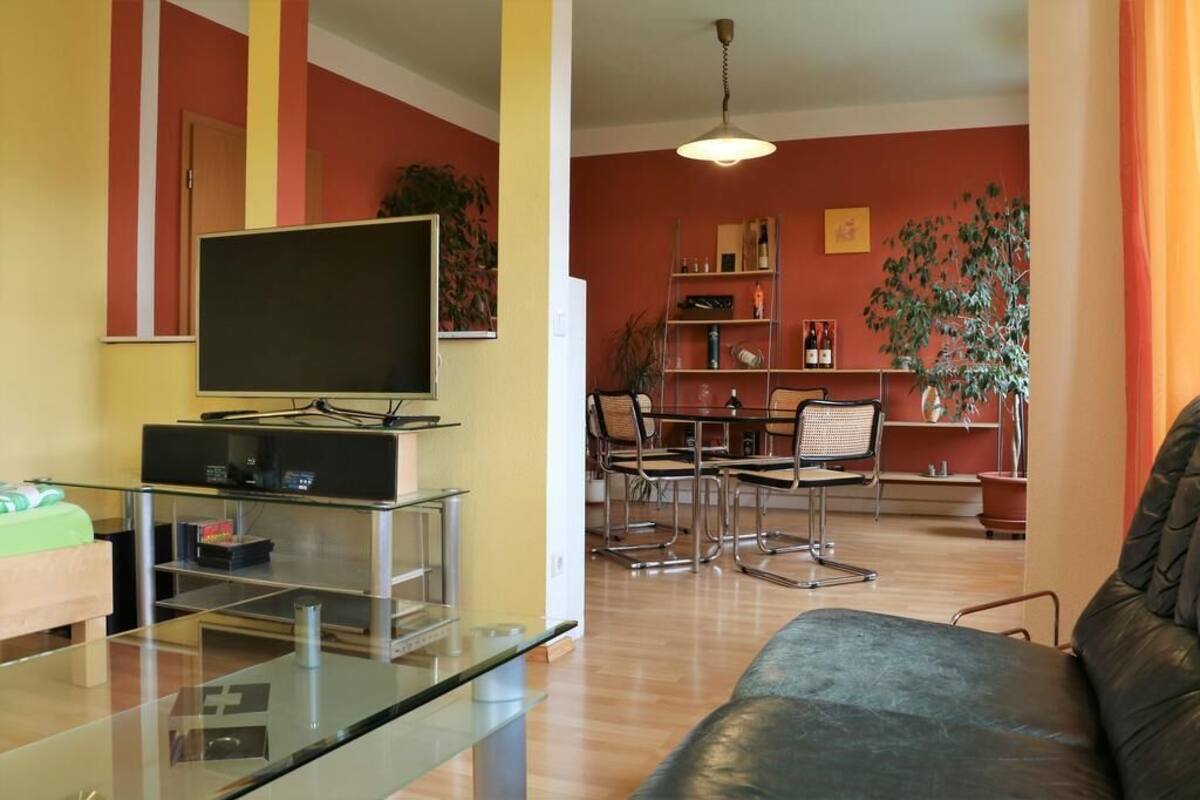 A living room with a flat-screen TV, glass coffee table, and a leather sofa. There is a dining area with a table and chairs, and a bookshelf with various items.