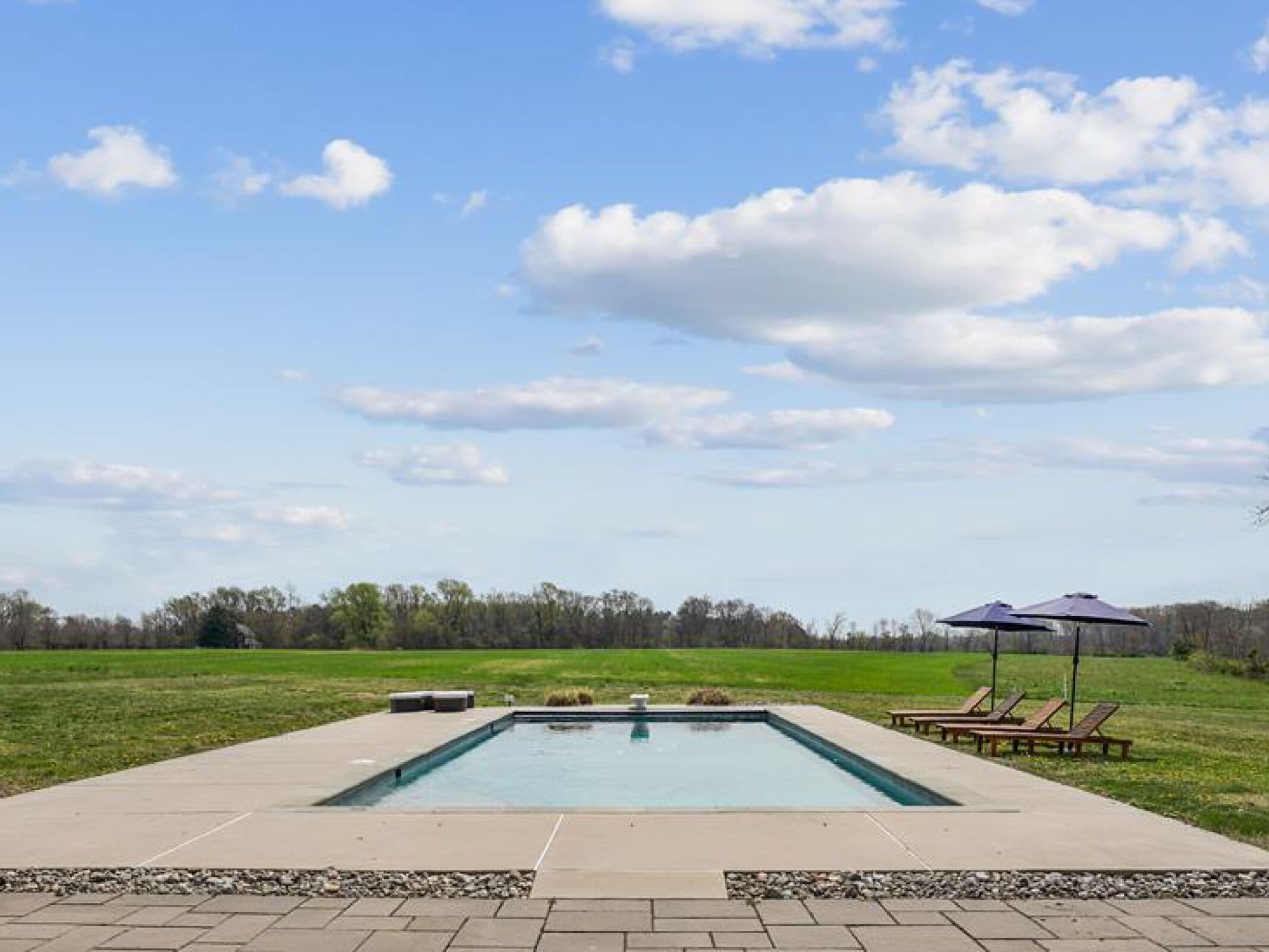 En rektangulär swimmingpool med en stenlagd poolterrass, omgiven av solstolar och ett picknickbord, med en gräsyta och träd i bakgrunden under en delvis molnig himmel.
